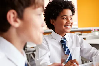 This image shows a photo of a child around 8-10 years old wearing a white shirt and blue striped tie as he sits in the school cafeteria smiling and laughing with his friends. 