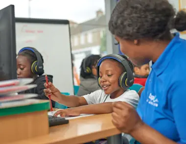 Children wearing headphones work on computers in a tuition centre while a tutor in a blue shirt assists one of them with their task.