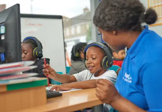Children wearing headphones work on computers in a tuition centre while a tutor in a blue shirt assists one of them with their task.