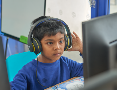 A young boy sits looking at a computer with headphones.