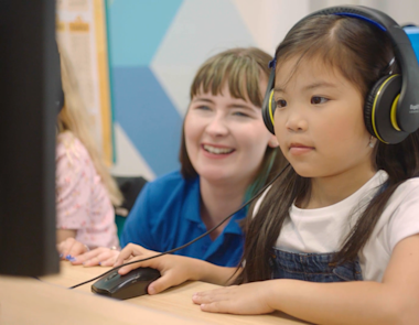 A young girl sits at a computer working with their tutor behind them assisting.